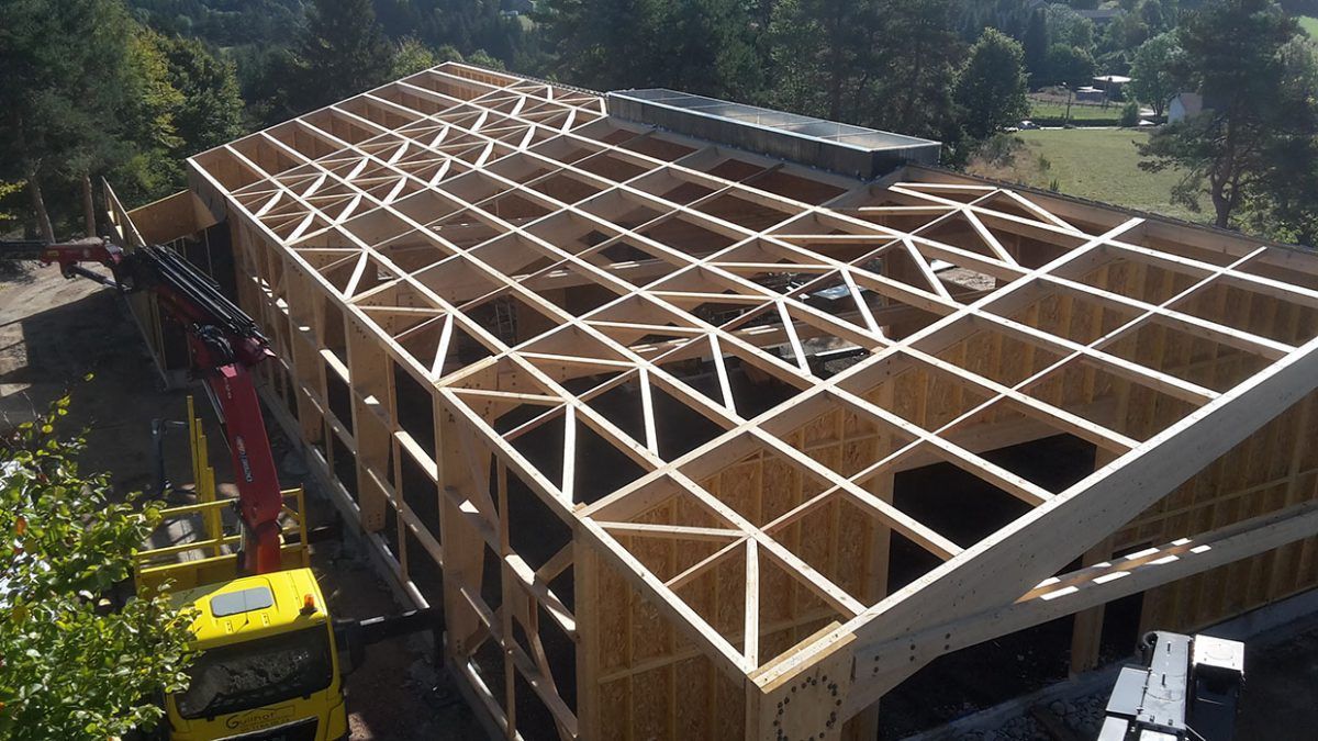 Bâtiment en bois en construction, charpente apparente, bras de grue jaune, lumière du jour.