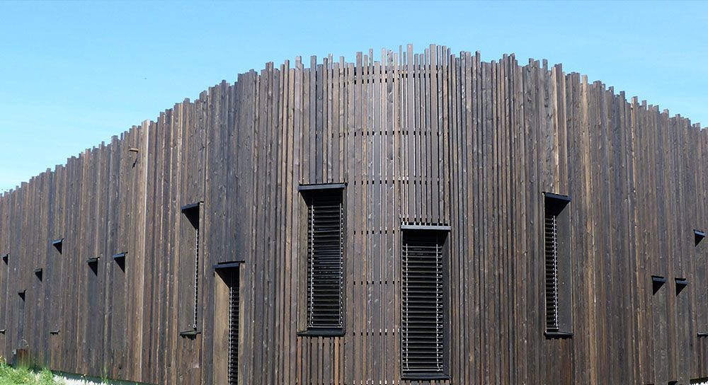 Coin d'un bâtiment aux lattes de bois verticales, brun foncé, et petites fenêtres carrées sous un ciel bleu.