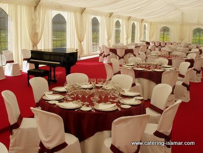 Elegante salón de banquetes con manteles rojos, fundas blancas para las sillas y un piano de cola bajo cortinas color crema.