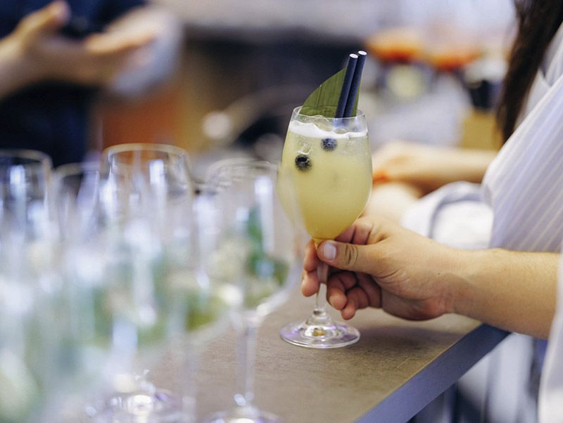 Una mano sostiene un cóctel amarillo alto con menta y frutos rojos en la barra de un bar.