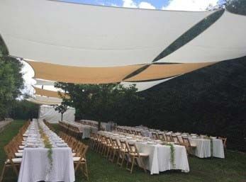 Recepción al aire libre con largas mesas de banquete cubiertas con manteles blancos bajo toldos extendidos en un jardín.