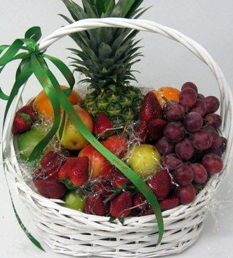 Cesta blanca con fruta variada: piña, uvas, fresas y manzanas, atada con una cinta verde.