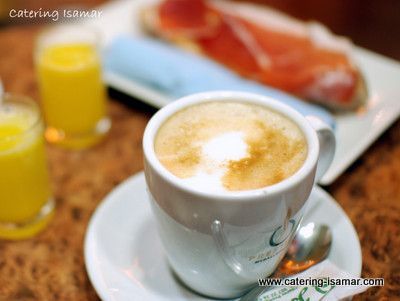 Un capuchino en una taza blanca junto a un zumo de naranja y bollería para el desayuno sobre una mesa.