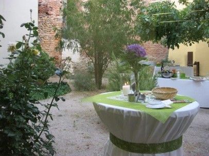 Mesa de patio con mantel verde, flores y vajilla junto a muros de ladrillo y árboles.