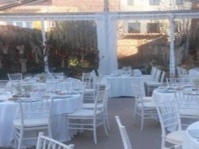 Montaje de banquete al aire libre con mesas y sillas blancas bajo una carpa transparente.