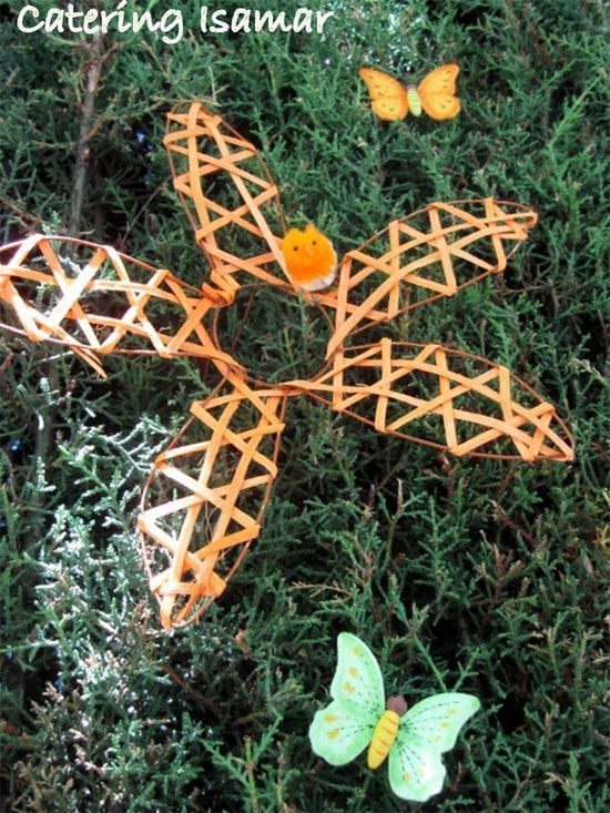 Adorno de estrella tejida de color naranja sobre follaje con dos mariposas, una amarilla y otra verde.