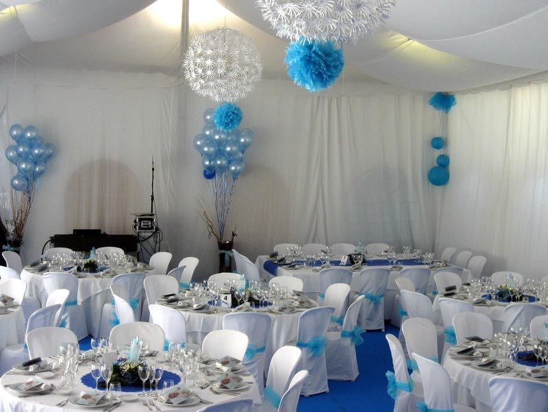 Carpa de banquete decorada con mesas redondas, fundas blancas para sillas y centros de mesa con globos azules y blancos.