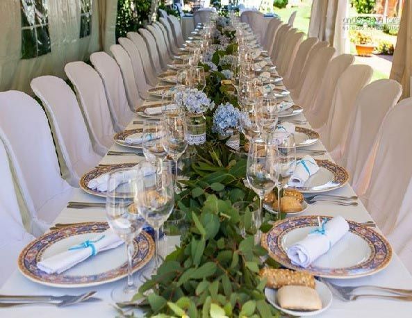 Mesa larga para banquetes al aire libre con sillas blancas, cristalería y centro de mesa con guirnalda floral verde.
