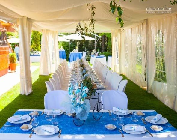 Mesa de banquete exterior decorada bajo un toldo blanco con detalles azules y vegetación colgante.