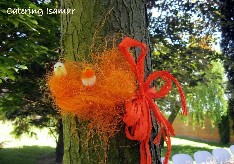 Adorno de pájaro de hilo naranja en el tronco de un árbol con cintas rojas en un jardín soleado.