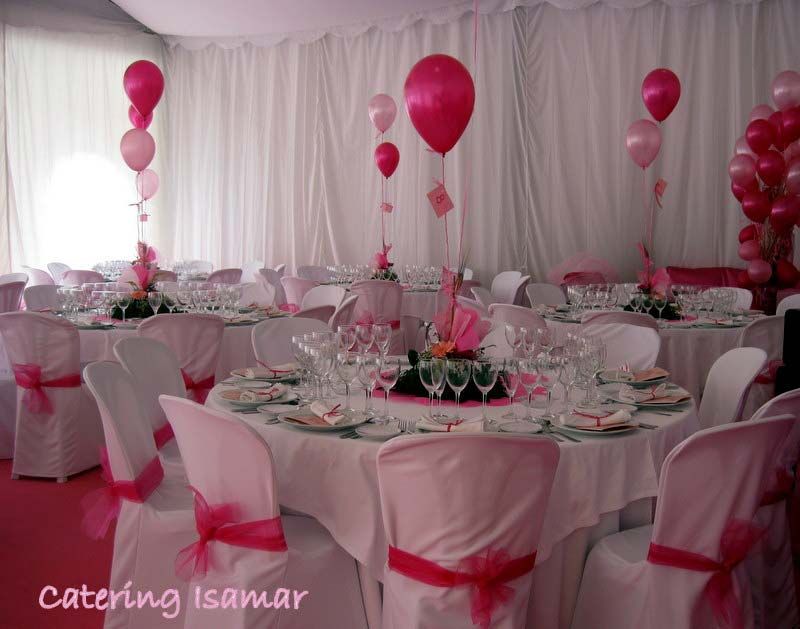 Salón de banquetes con temática rosa, mesas redondas, fundas blancas para las sillas y centros de mesa con globos rosas.