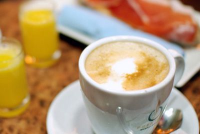 Una taza de capuchino con espuma sobre una mesa de desayuno, junto a un zumo de naranja y un plato de comida.