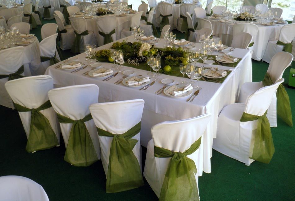 Mesa redonda de banquete con fundas blancas para las sillas y lazos verdes, preparada para un evento formal en interiores.