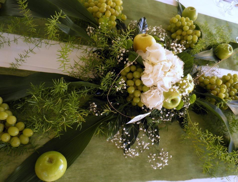 Centro de mesa floral verde y blanco con manzanas y racimos de uvas sobre un camino de mesa.
