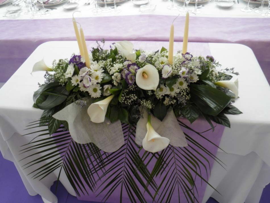 Centro de mesa floral con flores blancas y moradas sobre una mesa, adornado con velas altas y tela drapeada.