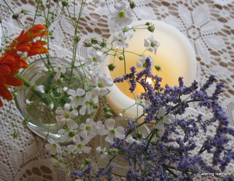 Velas en portavelas de cristal rodeadas de flores blancas, naranjas y moradas sobre un mantel de encaje.