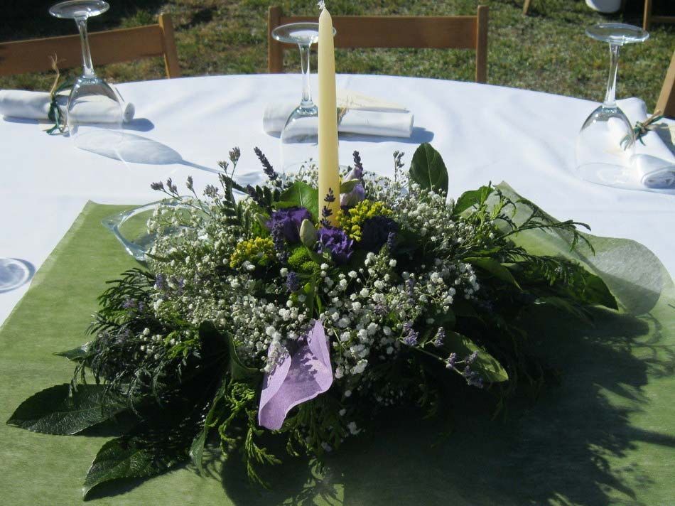 Centro de mesa floral iluminado con velas sobre una mesa blanca con camino de mesa verde y cinta morada.