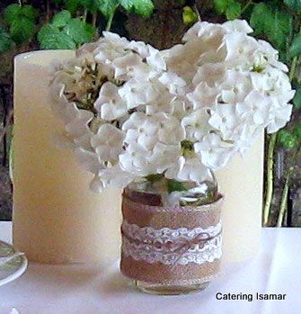 Ramo de flores blancas en un pequeño jarrón de cristal junto a una gran vela cilíndrica color crema sobre una mesa.