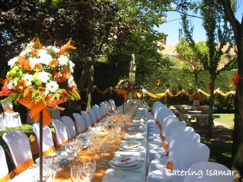 Mesa larga de banquete al aire libre con sillas blancas y centros de mesa florales en un jardín.