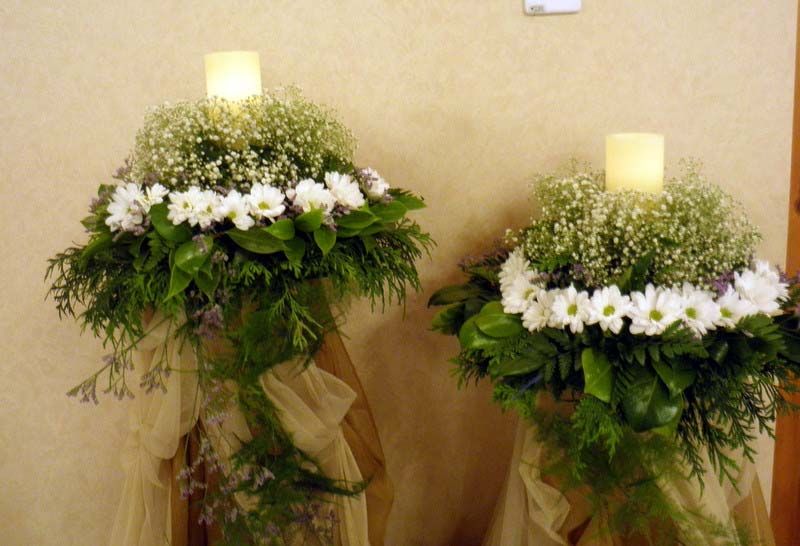 Dos arreglos florales con flores blancas y velas sobre soportes, contra una pared de color beige.