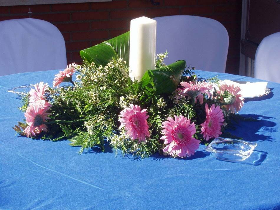 Centro de mesa con velas blancas, flores rosas y hojas verdes sobre un mantel azul.