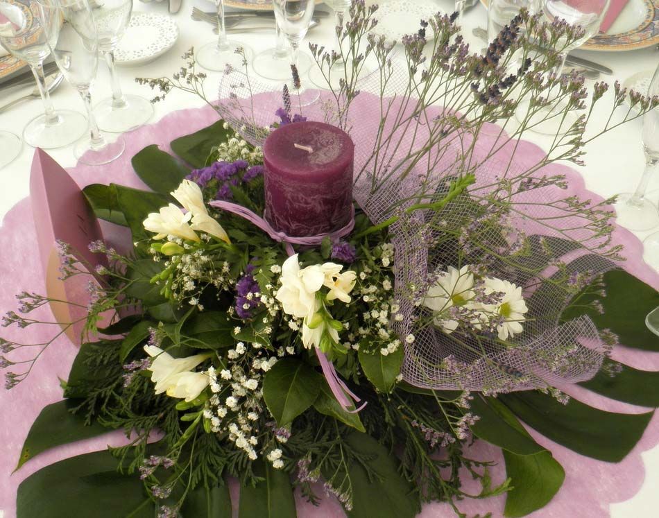 Centro de mesa floral morado y blanco con velas sobre un fondo rosa.