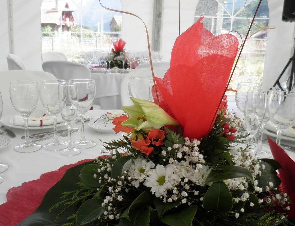 Elegante centro de mesa floral con flores rojas y blancas sobre una mesa puesta con copas de vino en una carpa para eventos luminosa.