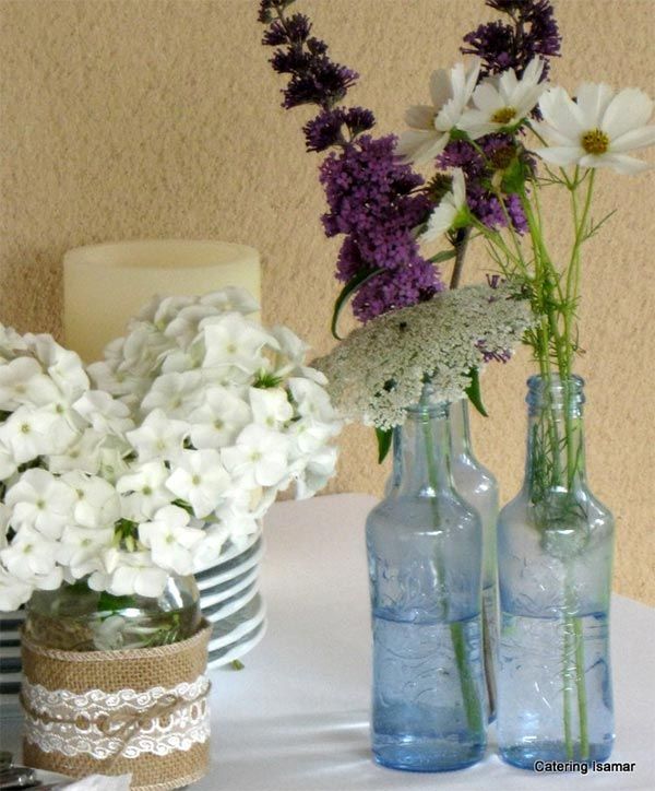 Variedad de flores blancas y moradas dispuestas en jarrones de cristal sobre un mantel blanco.
