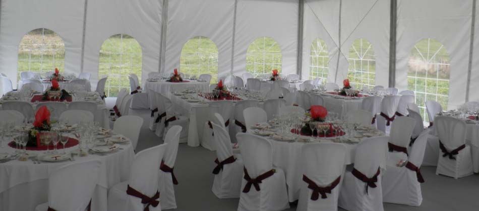 Carpa blanca para banquetes con mesas redondas y sillas vestidas de blanco, adornadas con centros de mesa florales rojos y decoraciones.
