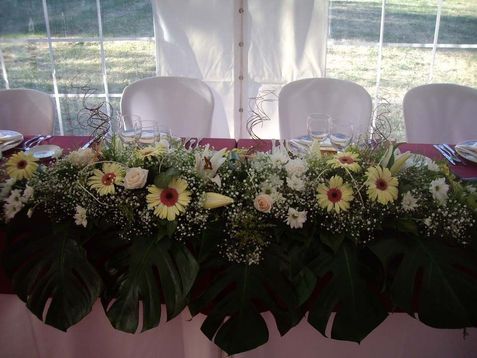 Arreglo floral en blanco, verde y rojo sobre una mesa de banquete decorada con sillas blancas detrás.