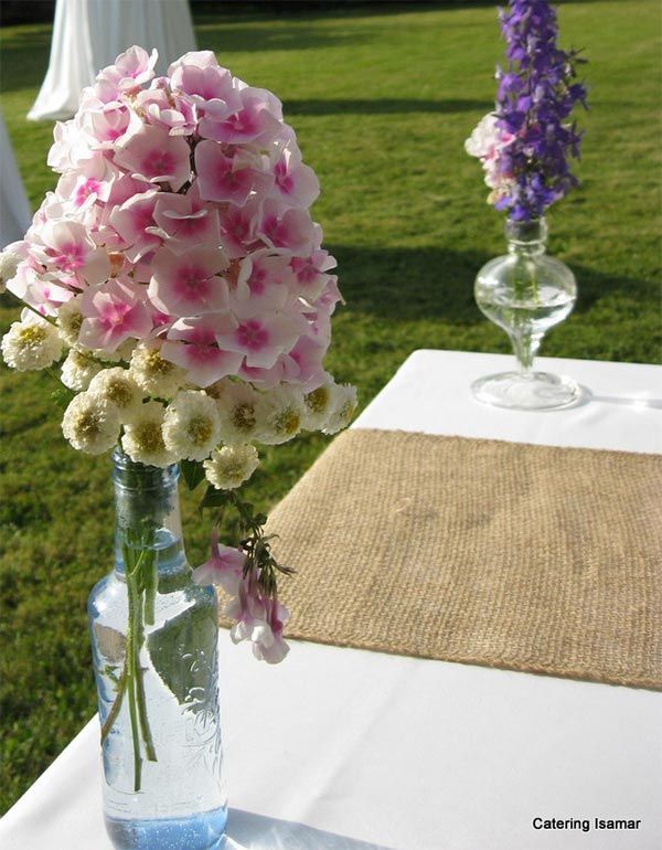 Centros de mesa florales sobre una mesa con camino de mesa de arpillera al aire libre en un césped soleado.
