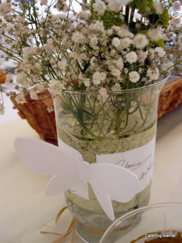 Pequeñas flores blancas dispuestas en un jarrón de cristal transparente con una mariposa blanca como adorno sobre una mesa.