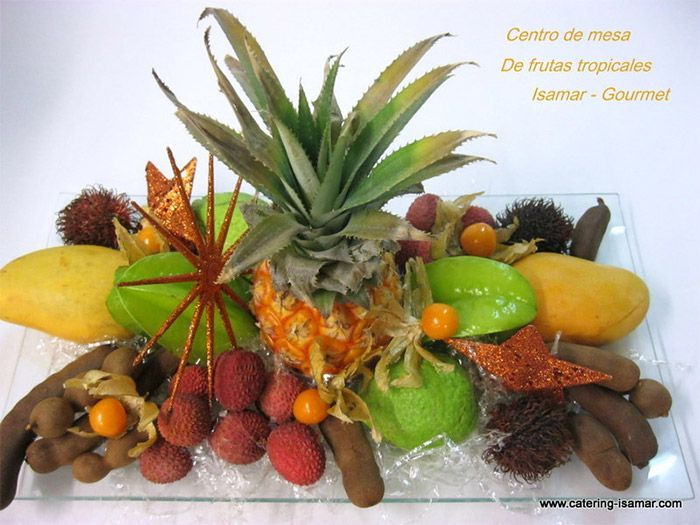 Centro de mesa frutal con piña, mangos, fresas, kiwi y carambola decorativa sobre una mesa.