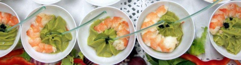 Cuatro tazas pequeñas de ensalada de camarones adornadas con aguacate sobre una mesa con frutos rojos y hojas verdes.