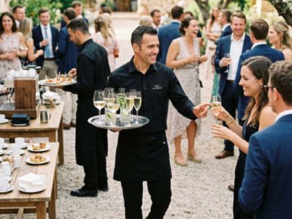 Un camarero sirve bebidas en una animada recepción al aire libre donde los invitados conversan.