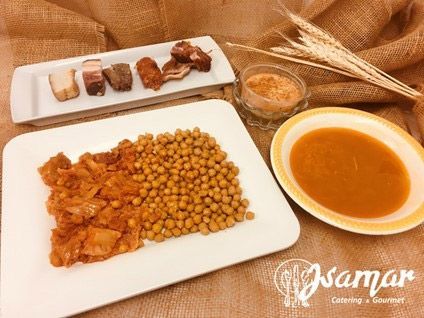 Vista cenital de una comida: garbanzos, pollo, pan, sopa y té en platos blancos con tallos de trigo sobre arpillera.
