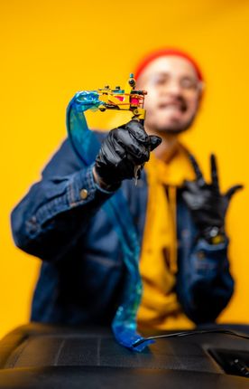 Una persona con una sudadera amarilla con capucha, una chaqueta vaquera azul y guantes negros sostiene una máquina de tatuar sobre un fondo amarillo brillante.