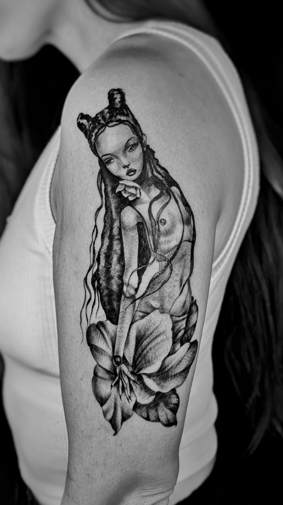 Un tatuaje en blanco y negro en la parte superior del brazo que muestra una figura ilustrada con cuernos, cabello largo y una base de orquídea.