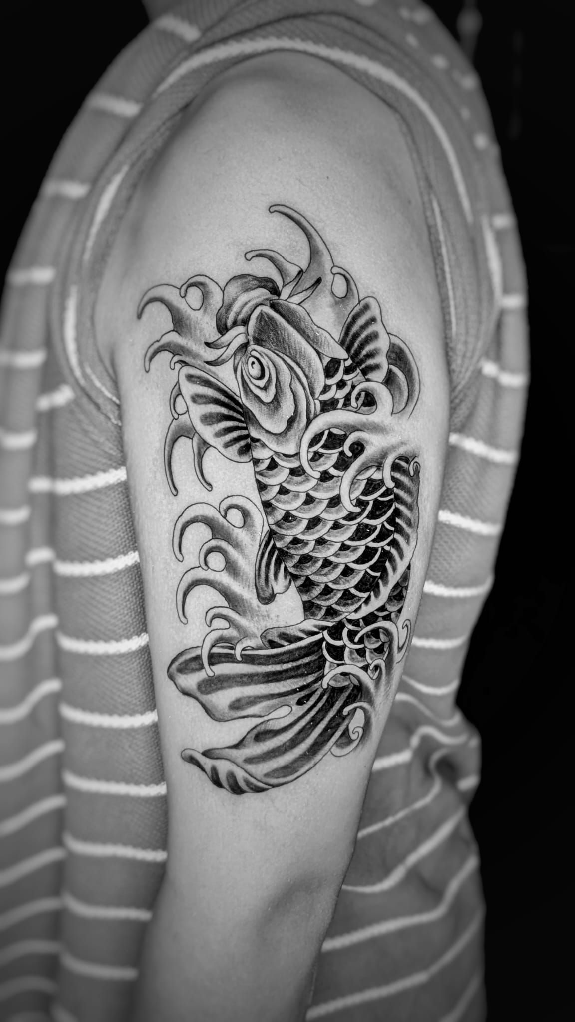 Un tatuaje en blanco y negro de un pez koi nadando entre olas de agua estilizadas en la parte superior del brazo de una persona.