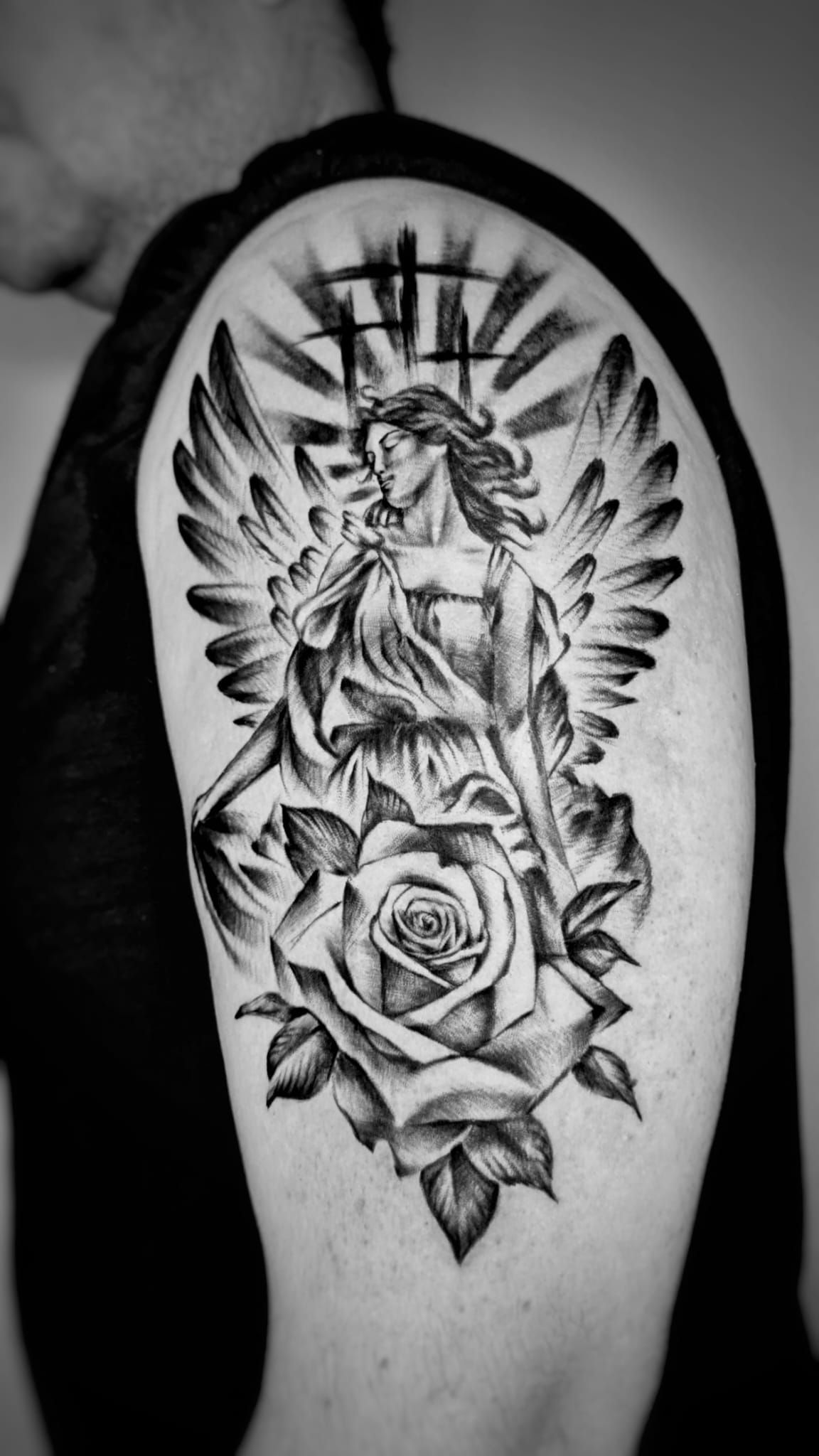 Un tatuaje en blanco y negro en la parte superior del brazo que representa a un ángel con las alas extendidas sobre una rosa grande y detallada.
