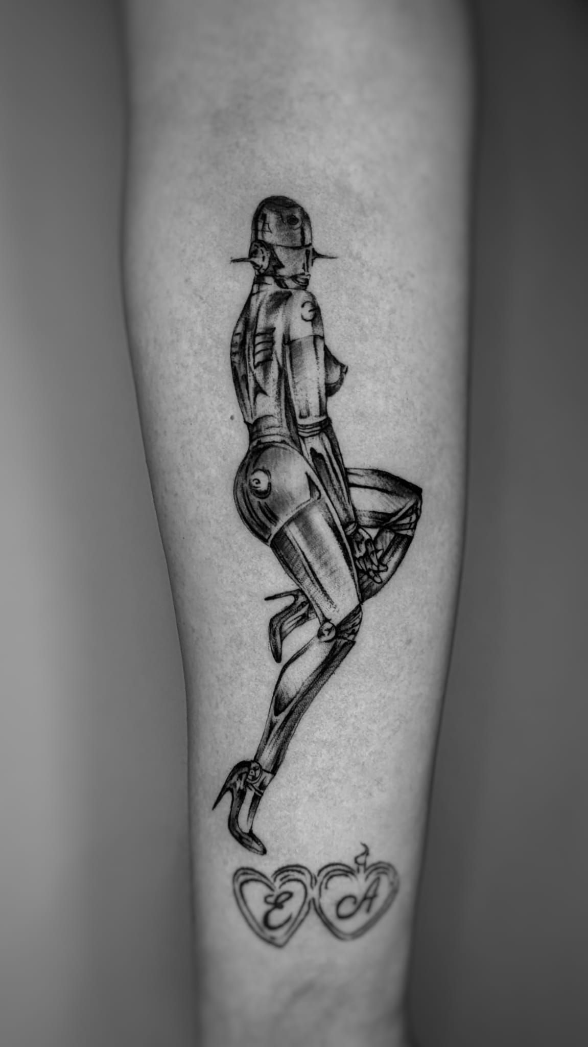 Un tatuaje de tinta negra en el antebrazo que representa una figura pin-up robótica estilizada sobre dos pequeñas formas de corazón que contienen iniciales.