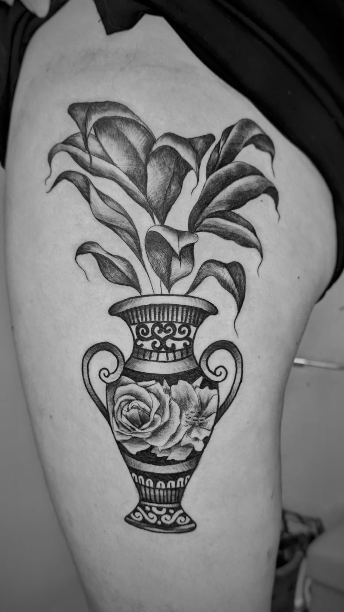 Un tatuaje en blanco y negro en la parte superior del muslo que representa una urna decorativa con plantas frondosas.