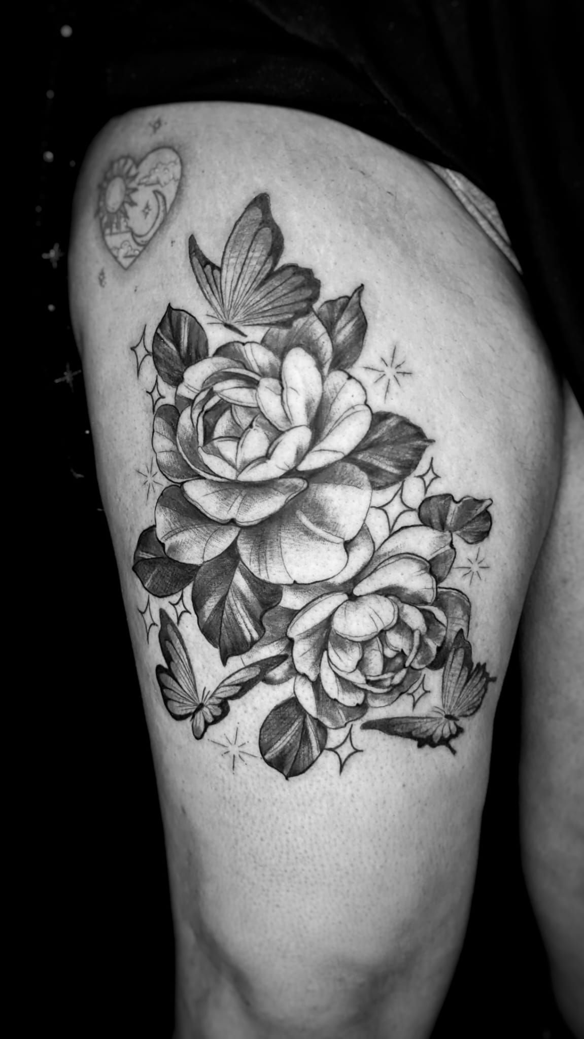 Tatuaje en blanco y negro en el muslo con dos rosas rodeadas de mariposas y pequeños detalles de estrellas brillantes.