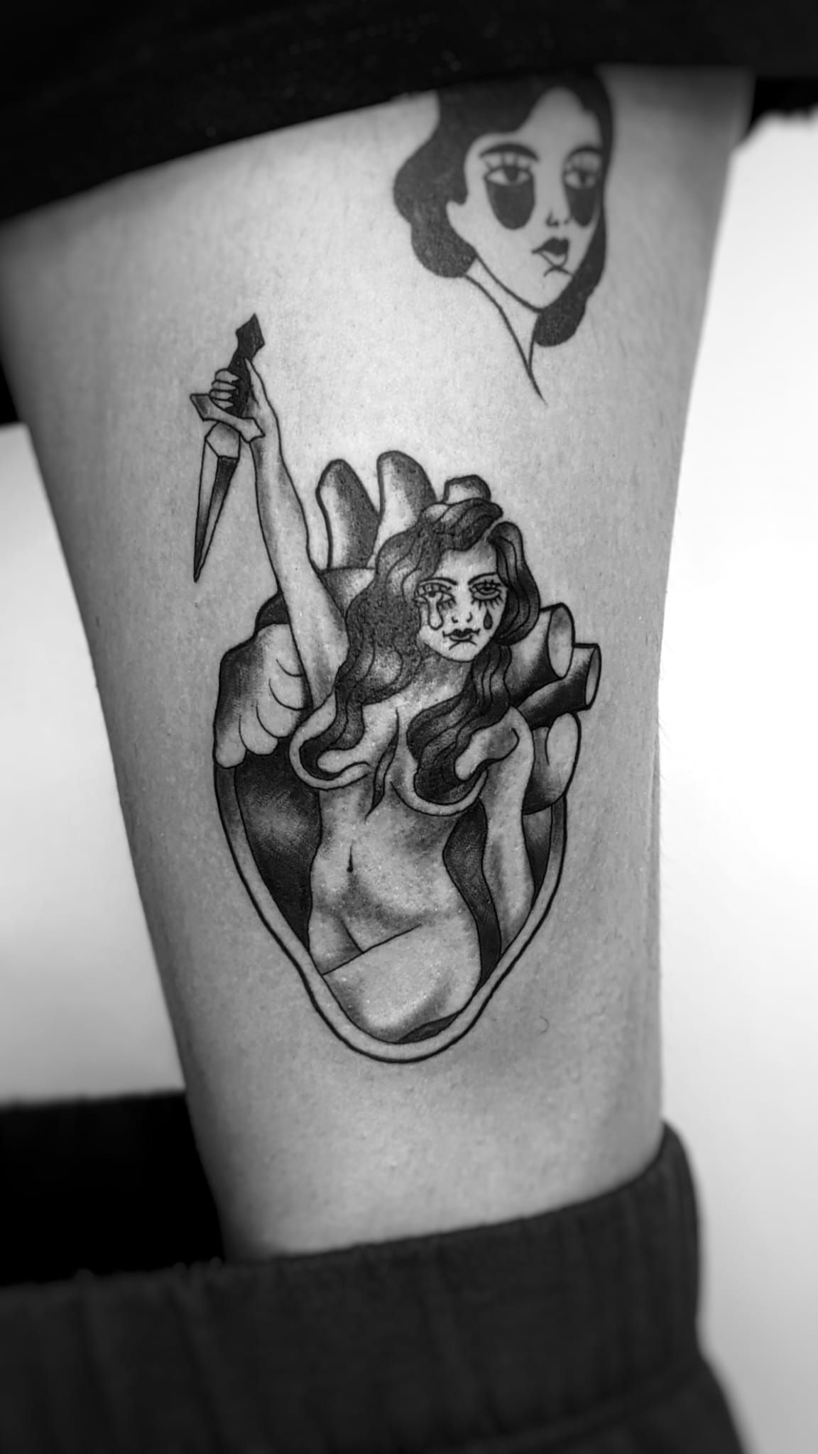 Tatuaje en blanco y negro sobre la piel que muestra una figura sosteniendo una daga dentro de un corazón, con un retrato tatuado encima.