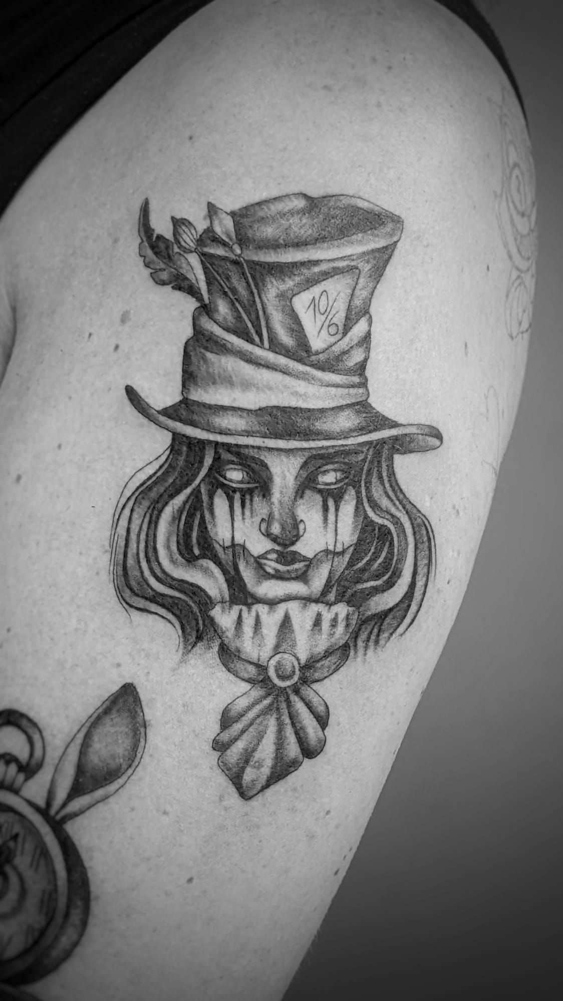 Un tatuaje en blanco y negro del Sombrerero Loco con un sombrero de copa con una tarjeta metida en el ala y una corbata con volantes.