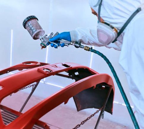 Un hombre está pintando con aerosol el parachoques de un coche rojo en un garaje.