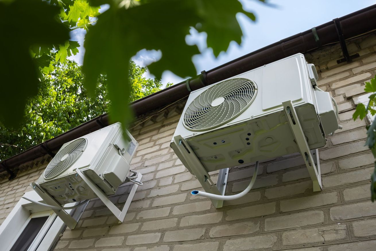 Deux unités de climatisation extérieures fixées sur un mur de briques et des feuilles vertes.