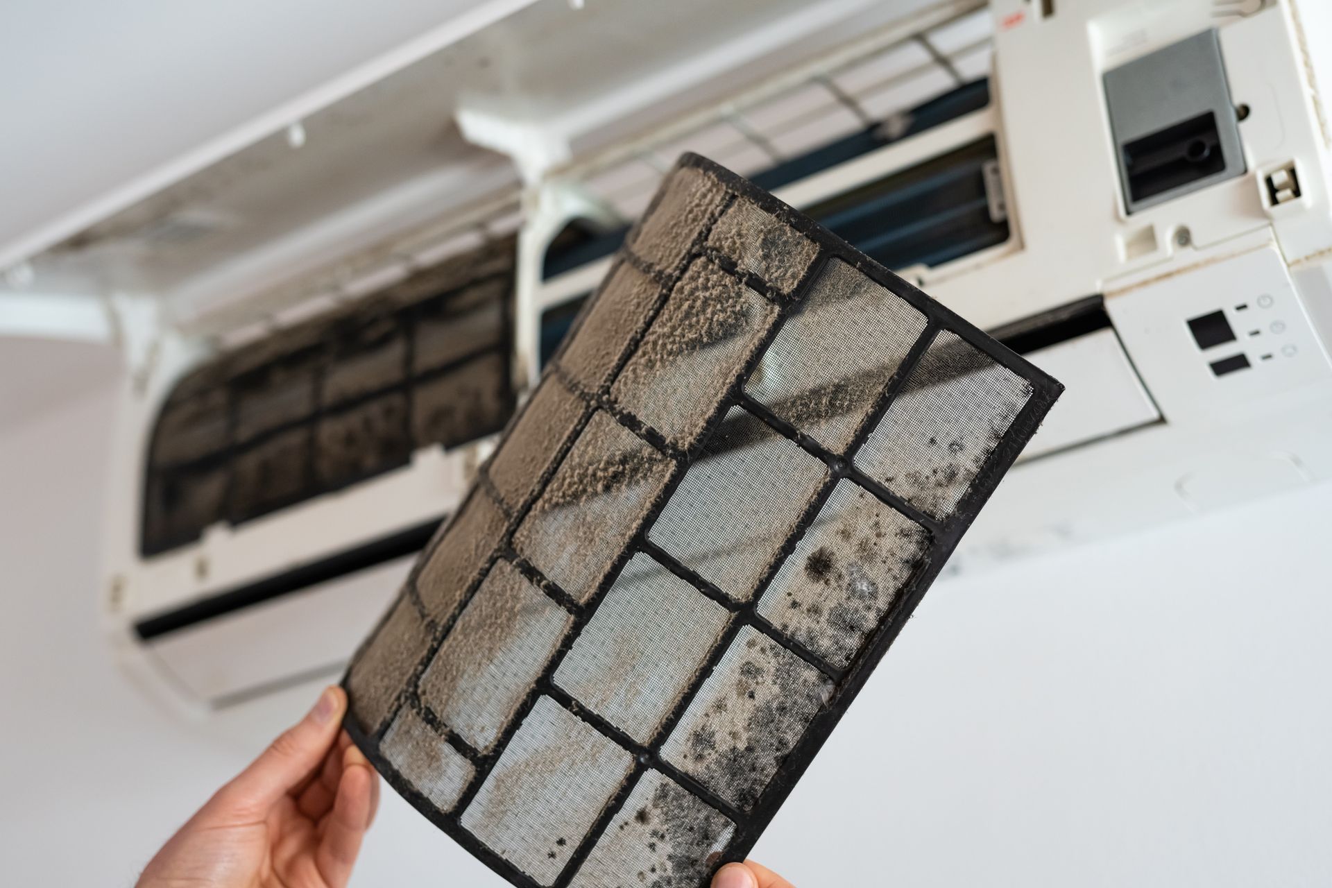 Une main tenant un filtre de climatiseur encrassé devant une unité de climatisation intérieure ouverte.