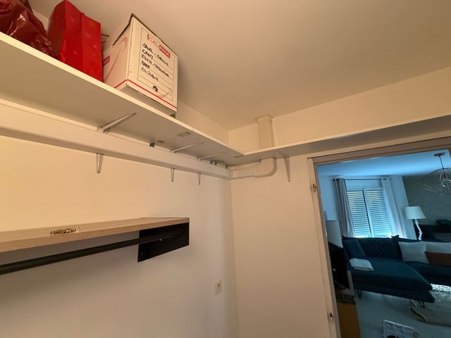 Petite pièce de rangement blanche avec des étagères, des cartons, un câblage caché et une porte donnant sur un salon éclairé en bleu.