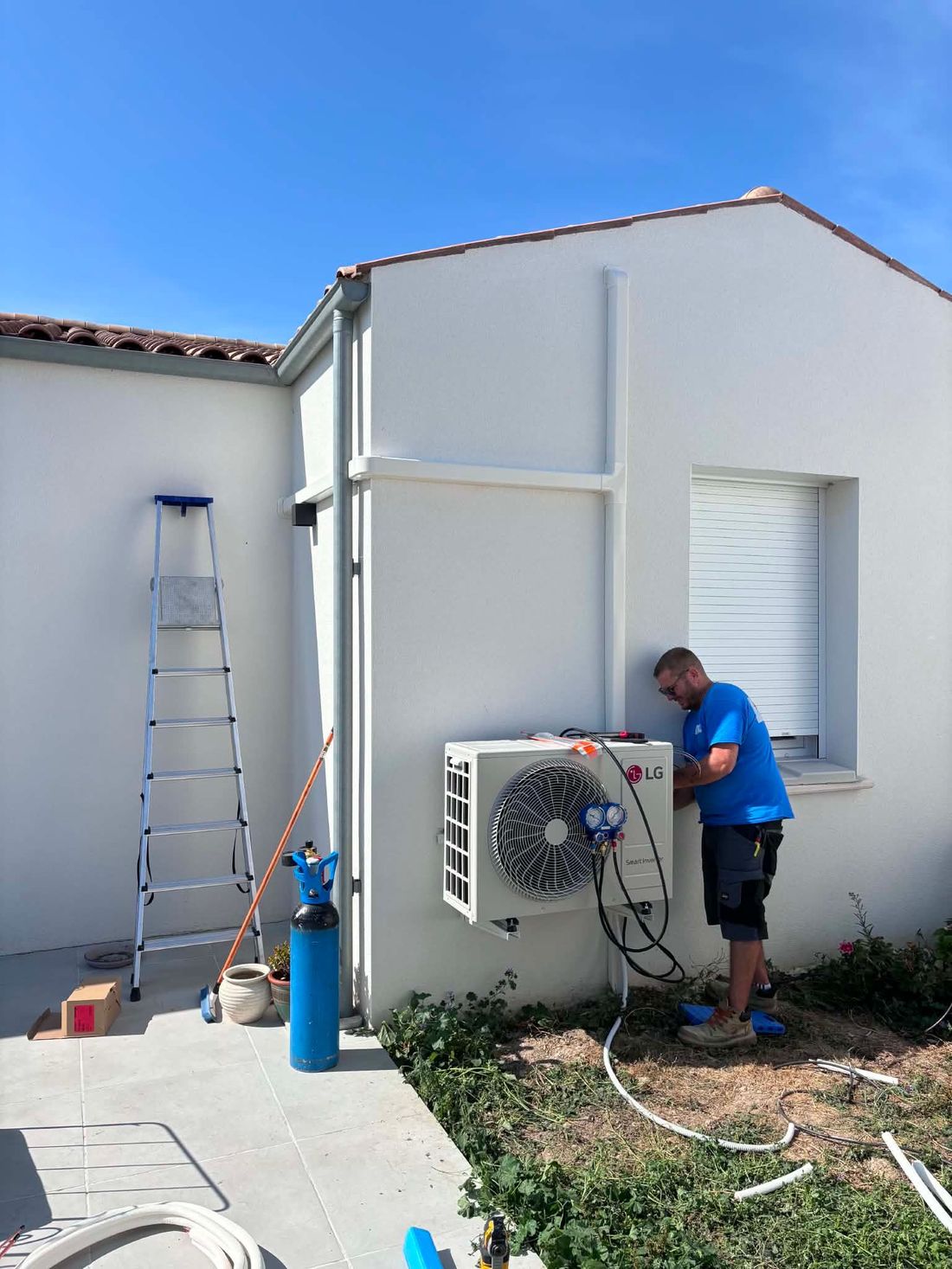 Un technicien travaille sur une unité de climatisation extérieure, près d’une maison blanche en stuc, avec une échelle et des outils à proximité.